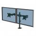 In the photo, two sleek and modernly designed monitor arms are attached to a desk. The arms are adjustable and are holding two computer monitors in a side-by-side, dual configuration. The arms have a sleek silver finish and are connected to the desk with a sturdy clamp.