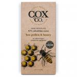 Cox & Co. Bee Pollen & Honey chocolate bar rests on a plain surface. The wrapper mixes warm yellow and deep brown with a neat logo and bee illustration. A partly unwrapped dark chocolate square shows golden pollen specks and a glossy honey drizzle. The photo uses soft studio lighting. Neutral tones.