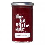 Six glass jars of The Bit on the Side strawberry jam sit in neat rows, showing deep red fruit spread through clear glass. Each jar bears a white label with the brand name and a stylized strawberry illustration; silver screw lids catch the light, and a cardboard tray frames the group. Orderly display