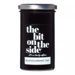Six glass jars of The Bit on the Side Blackcurrant Jam sit in tidy rows on a wooden board. Deep purple jam gleams through clear glass, black lids display branded labels in white and purple. Scattered blackcurrants and a silver spoon add texture and contrast.