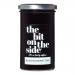Six glass jars of The Bit on the Side Blackcurrant Jam sit in tidy rows on a wooden board. Deep purple jam gleams through clear glass, black lids display branded labels in white and purple. Scattered blackcurrants and a silver spoon add texture and contrast.