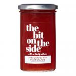 Six clear glass jars of pink-orange marmalade sit in neat rows. Each jar wears a cream label with pink accents and a gold lid; translucent, pulpy marmalade shows citrus segments. Grapefruit slices and elderflower sprigs rest on a light wooden surface under soft natural light.
