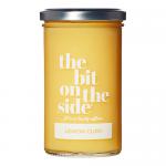 A cluster of six glass jars of lemon curd sits on a light tabletop. Each jar displays a pale yellow, glossy curd visible through clear glass, topped by a cream lid. Labels show simple lemon illustrations, bold brand text and subtle decorative accents.