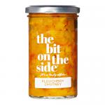 Six glass jars of Ploughboy chutney sit neatly in a cardboard tray; deep amber chutney glows through clear glass. Cream labels feature bold blue logo 'The Bit on the Side', rustic typography and small vegetable illustrations. Metal lids reflect studio lighting. Soft shadows on pale wood. Dewy beads.
