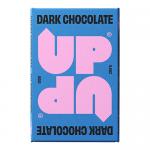 A dark chocolate bar and its wrapper sit on a neutral surface: the bar displays glossy, deep-brown squares with clean edges and a subtle sheen; the wrapper shows bold lettering and dark tones, with a torn opening revealing the bar inside. Soft diffuse light casts a warm shadow and a torn foil peeks.
