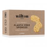 A twin pack of Wilton London eco sponges sits on a plain surface. Two compact rectangular sponges show a soft beige cellulose side and a textured green scouring layer. A recyclable paper band wraps them, displaying minimalist branding and simple product text. Soft studio lighting casts shadows. Dim.