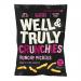 Photo shows a 100g Well & Truly Crunchies Punchy Pickle bag beside a neat pile of fourteen smaller Well & Truly crisps packets. Vibrant green and yellow wrappers display pickle graphics and bold white branding. Several open packs reveal ridged, golden chips on a pale surface.