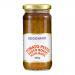 Photo shows a glass jar of Seggiano tomato pesto, the deep red-orange spread visible through clear glass. A cream label with brand text and tomato illustration wraps the jar. Glossy metal lid reflects light. Spoon beside jar reveals chunky, herb-flecked texture. Soft light reveals tiny tomato seeds.