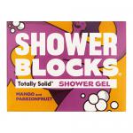 Six vibrant mango-orange solid shower gel blocks sit arranged on a white ceramic tray. Tropical fruit illustrations and sliced mangoes and passionfruit cluster nearby. Soft natural light casts gentle shadows; a folded towel and green leaves add lifestyle context. Subtle condensation dots the surface.
