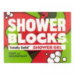 Six solid shower blocks sit arranged on a wooden tray and in open cardboard box, alternating mint-green and coral-pink bars. Fresh mint leaves and halved grapefruit rest beside them on wet ceramic tile, with soft natural light and water droplets highlighting textures. Soft reflections on glossy edges