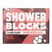 Six deep-red solid shower gel blocks sit stacked on a wooden tray, their glossy surfaces dotted with water droplets. A black cherry slice and green leaves rest nearby, while soft natural light highlights textured ceramic tiles and a chrome showerhead in the blurred background. Subtle sheen glimmers.