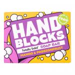 Photo shows six hand-sized soap blocks arranged on a wooden tray atop a folded linen cloth. Bars display swirls of bright mango orange and deep passionfruit purple. Fresh mango and passionfruit halves, glossy green leaves and soap curls sit nearby under soft daylight.