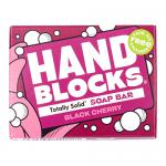 A lifestyle photo shows six hand-block soap bars in rich black-cherry hues, each bearing subtle carved patterns and slightly rustic edges. Bars sit stacked and fanned on a wooden board with scattered fresh cherries and green leaves, soft natural light and gentle shadows revealing texture. Close-ups.