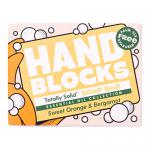 Six hand-shaped soap blocks in warm orange tones sit on a wooden tray, their surfaces showing subtle marbling and pressed botanical textures. A sliced sweet orange and bergamot fruit rest nearby as soft natural light casts gentle shadows across a rustic lifestyle setting, with scattered citrus peel.