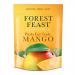 A clear front pouch displays bright orange dried mango slices alongside glossy almonds, pumpkin seeds and sunflower kernels. A simple matte label at top shows the brand name and product title in green lettering, with nutritional icons and a small weight indicator. The backdrop is a pale wood surface.