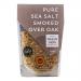A clear glass jar holds coarse, grey-brown smoked sea salt beneath a light wooden lid. A cream label displays the Halen Môn logo and black text. Visible crystals show smoky flecks and varied textures, creating a rustic, handcrafted look.