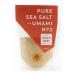 Photo shows a compact jar of Anglesey sea salt labeled Halen Môn Umami. Clear glass reveals pale, crystalline flakes; a simple label with logo and text wraps the jar. A black screw cap tops the jar, which sits on a neutral surface with soft lighting.