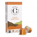 A rectangular box stands upright with bright orange and cream graphics and a prominent logo. Several single-serve coffee pods sit beside it, showing metallic foil lids and ribbed plastic cups. Small ingredient and label text runs along the box sides.