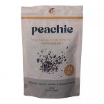 A clear glass jar with a peach-toned label sits against a neutral background. The lid gleams; inside a crunchy mix of hazelnut pieces, cacao nibs, dried cherry bits and mixed seeds creates a textured, earthy palette of browns, deep reds and cream hues, with visible crumbs and flakes. Soft highlights.