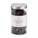 A clear plastic pouch displays irregular, glossy black fungus pieces—thin, ruffled, ear-like shapes with wrinkled, dark surfaces. A simple white label shows the Emma Basic brand and product details; the pouch rests against a plain, light background. Soft light reveals glossy sheen and delicate edges