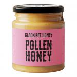 Six glass jars sit in a neat row, filled with amber honey and flecks of pollen. Labels feature a stylized black bee and British flag motifs in black and gold. Metal screw lids reflect light. A simple white background emphasizes the warm, glossy texture of the honey. Soft shadows add warmth and depth.