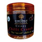 Six 100g clear glass jars of crispy chilli crunch sit arranged in two neat rows; the jars reveal red-brown chili and crunchy coconut pieces, each capped with a metallic lid and a cream label bearing bold brand text and a small coconut illustration against a plain white background with soft lighting