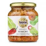 Six 350g jars of Biona Organic kimchi sit in two neat rows, their clear glass revealing vibrant red-orange fermented cabbage and visible chili flecks. Each jar wears a white label with green Biona branding and a black lid; condensation and textured cabbage pieces add visual freshness. Bright labels.