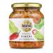 Six 350g jars of Biona Organic kimchi sit in two neat rows, their clear glass revealing vibrant red-orange fermented cabbage and visible chili flecks. Each jar wears a white label with green Biona branding and a black lid; condensation and textured cabbage pieces add visual freshness. Bright labels.