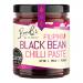 A glass jar with a deep brown-black paste, flecked with red chili seeds and glossy oil, sits against a plain white background. The label shows bold black and gold lettering with the brand logo, ingredient icons, and a red chili illustration wrapping part of the jar. A gold lid gleams beneath lights.
