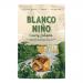 A bright bag shows the Blanco Niño logo atop a creamy jalapeño graphic. Golden, triangular tortilla chips spill from the package, speckled with seasoning. Fresh jalapeño slices and a swirl of pale green dip accent the vibrant, snack-focused composition. Soft studio light and pale teal backdrop glow.