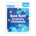 Six Brave Roasted Chickpeas Sea Salt sharing bags sit clustered on a neutral surface. Each matte pouch shows the Brave logo, product name and a clear window revealing golden-brown chickpeas. A small heap of roasted chickpeas and scattered sea-salt flakes lie in front. Soft shadows fall across scene.