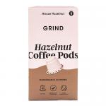 Photograph shows a compact box of Grind Coffee pods featuring soft beige and warm brown tones with stylized hazelnut imagery and subtle green compostable icons. Packaging displays clean white typography and weight markings. Ten glossy metal-look pods peek from open flap on a neutral kitchen counter.