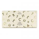 A glossy rectangular box sits centered, deep brown lid embossed with gold lettering and a delicate border. Twelve round chocolate truffles rest in a molded tray, each dusted or coated differently — matte cacao, shiny ganache, and sprinkled textures — with a ribbon accent at one corner. Subtle sheen.