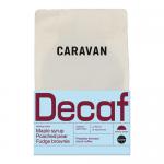 A matte brown 200g bag of Caravan Coffee Roasters decaf beans stands upright, showing a minimalist label with the Caravan logo and product name. Loose dark beans lie beside the bag on a light wooden surface under soft natural light.