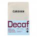 A matte brown 200g bag of Caravan Coffee Roasters decaf beans stands upright, showing a minimalist label with the Caravan logo and product name. Loose dark beans lie beside the bag on a light wooden surface under soft natural light.