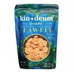 A matte white pouch displays Kin + Deum branding beside green waves; a clear window reveals dark-green, irregular seaweed-rimmed rice crisps. Crisps appear thin, bubbled and pale, scattered on a wooden board beside loose flakes of dried seaweed. Foil accents catch light; tiny salt crystals glinting.