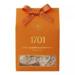 A rectangular gift box sits on a neutral surface, its lid displaying glossy photos of honey-drizzled nougat studded with whole almonds and chopped dates, dusted with crushed cardamom. Soft gold lettering and a subtle patterned border frame the image. A small translucent window shows neat bite slices.