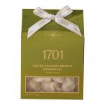 A compact rectangular box labeled Salted Caramel Brittle and Pistachio Honey Nougat features warm caramel tones and pistachio-green accents. A clear window reveals glossy amber caramel shards and creamy nougat studded with green pistachio pieces against a clean white backdrop.