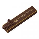 Photograph shows a glossy Belgian chocolate–coated honey nougat bar partly sliced to reveal a soft, ivory, honey-colored interior studded with roasted nuts. The bar sits on a crinkled golden wrapper embossed with a simple confectionery logo against a dark backdrop.
