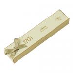 A wrapped roasted-almond honey nougat bar rests on a neutral backdrop. The packaging shows warm amber and cream hues, stylized almonds and honey drips. A clear window reveals dense ivory nougat studded with toasted almond pieces and a glossy honey sheen. Soft wrapper edges glint under soft lighting.