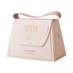 A small handbag-shaped box in warm cream and honey tones sits against a white background. A clear window reveals pale nougat studded with macadamia halves and amber honey swirls; gold lettering and a ribbon handle finish the packaging.