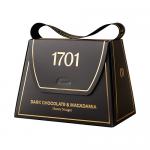 A small handbag-shaped box rests on a neutral background, its glossy dark-brown packaging edged with gold foil. Inside, irregular dark chocolate pieces studded with macadamia halves surround pale honey nougat; scattered nuts and a honey glaze catch the light.