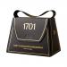 A small handbag-shaped box rests on a neutral background, its glossy dark-brown packaging edged with gold foil. Inside, irregular dark chocolate pieces studded with macadamia halves surround pale honey nougat; scattered nuts and a honey glaze catch the light.