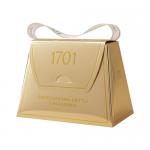 A handbag-shaped tin with a glossy caramel-brown exterior, embossed lettering and a tied ribbon. The lid opens to reveal golden salted caramel brittle and pale macadamia honey nougat pieces studded with glossy nuts, set against a crinkled paper liner.