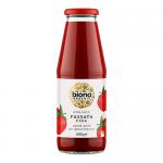 A clear glass jar of smooth, vibrant red tomato purée stands upright on a neutral background. A white-and-green label wraps the jar, showing the Biona logo and botanical illustrations of tomatoes and leaves. A ridged metal screw lid seals the top.