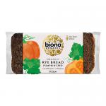 The image shows a dense round rye loaf wrapped in clear packaging with the Biona label; a crackled dark crust exposes a moist, speckled interior heavy with green pumpkin seeds. A few loose seeds scatter on a wooden board beside a serrated knife and natural linen cloth. Warm light reveals crumb grain