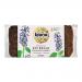 A rustic dark rye loaf rests in clear plastic packaging with a brown kraft label and Biona Bakery logo. Dense, speckled slices reveal chia and flax seeds throughout. The crust is deep brown and slightly cracked, and seeds scatter across the surface visible through the bag.