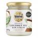 A set of six glass jars of Biona Organic Raw Virgin Coconut Oil sits on a neutral background. Each clear jar shows white solid coconut oil and carries a green-and-white label with the Biona logo, product name, organic certifications, and a screw-top lid. Lined up in two neat rows lit with soft light.