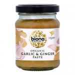 A cluster of six clear glass jars sits in a plain cardboard tray, each filled with pale beige garlic-ginger paste. Green lids cap uniform white labels bearing the Biona logo and organic markings, plus small illustrations of garlic bulbs and ginger roots. Soft light reveals faint ridges soft shadows.