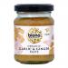 A cluster of six clear glass jars sits in a plain cardboard tray, each filled with pale beige garlic-ginger paste. Green lids cap uniform white labels bearing the Biona logo and organic markings, plus small illustrations of garlic bulbs and ginger roots. Soft light reveals faint ridges soft shadows.